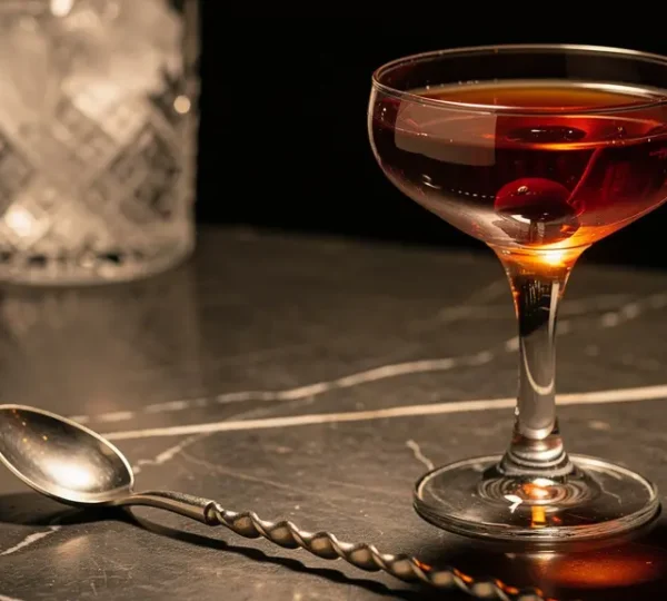 A perfectly stirred Manhattan cocktail in a coupe glass with jewel-like ruby clarity, a bar spoon resting beside it on a dark marble surface.
