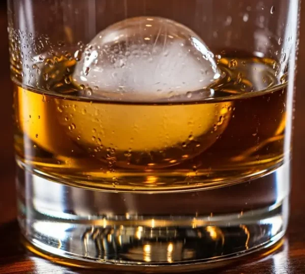 Cinematic close-up of a heavy-bottomed crystal Old Fashioned glass containing a whiskey cocktail with a large clear ice sphere, set on a dark wooden surface.