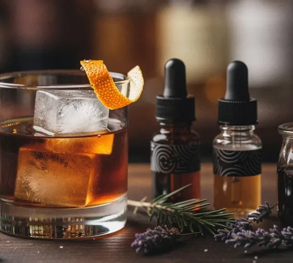 An Old Fashioned on a bar top with unlabeled bitters bottles, a small jar of syrup, and citrus peels arranged with generous negative space.